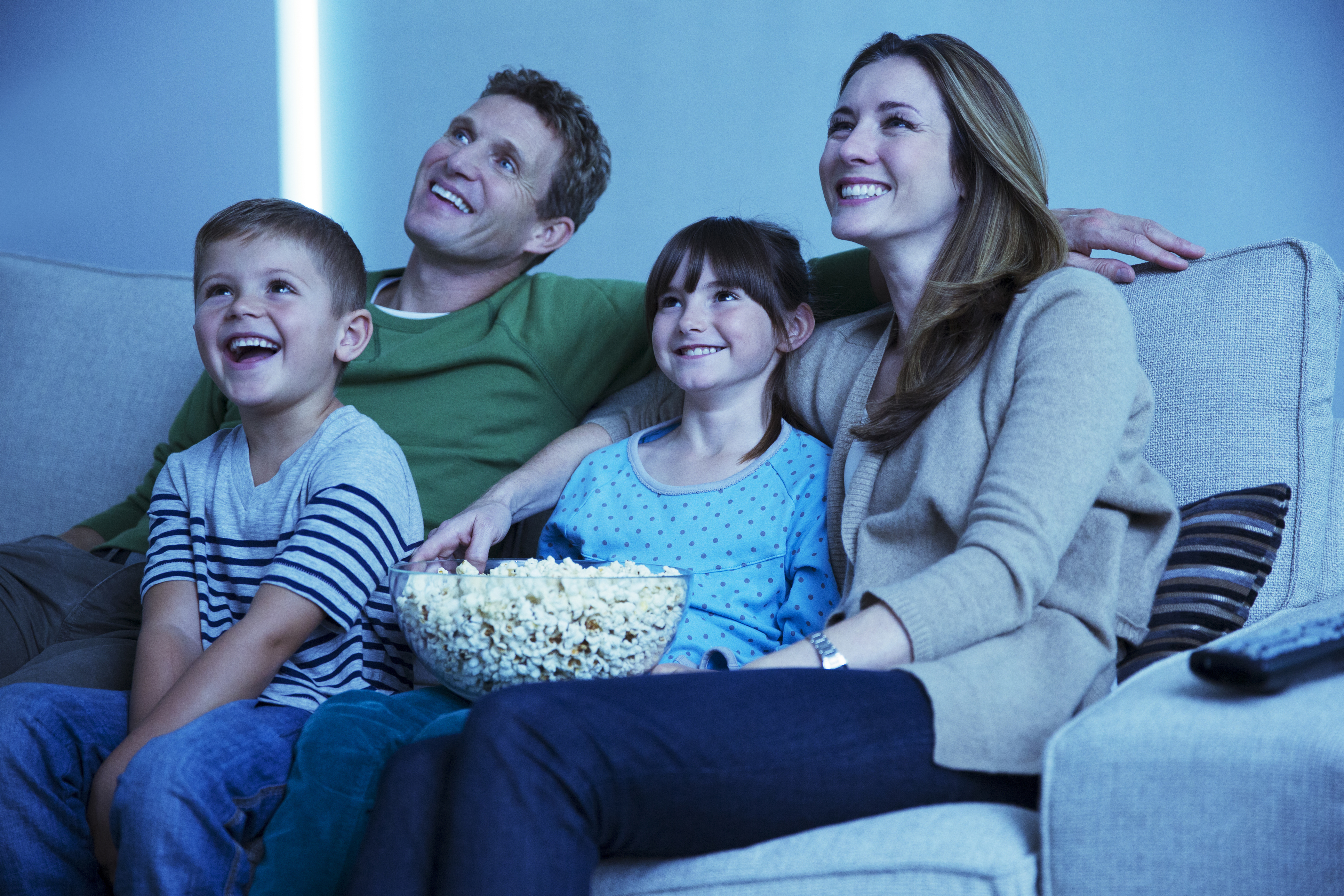 A family of four laughing on a sofa while watching entertainment and eating popcorn 