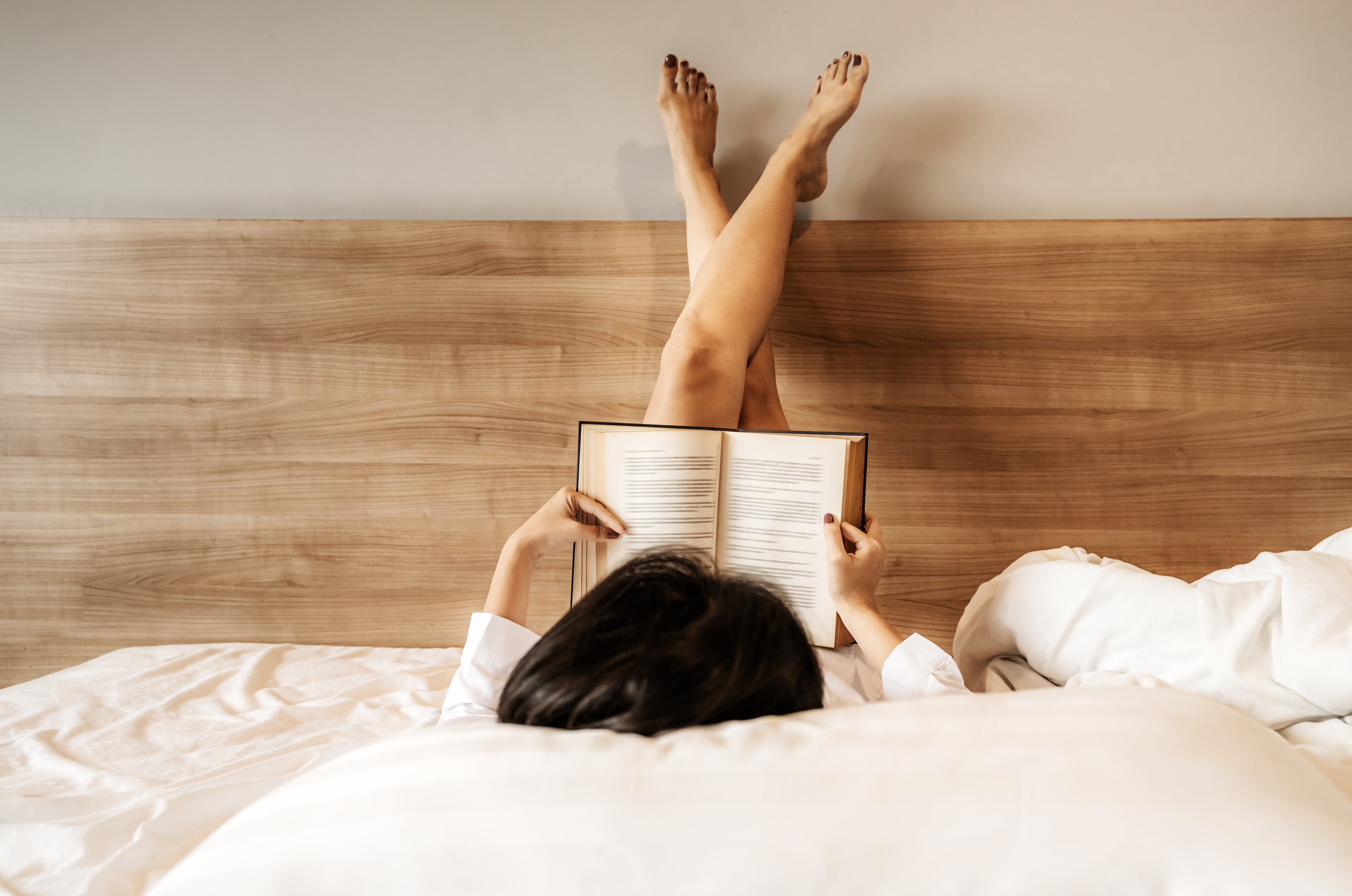 : A woman with her feet up on a headboard reading a book in bed 