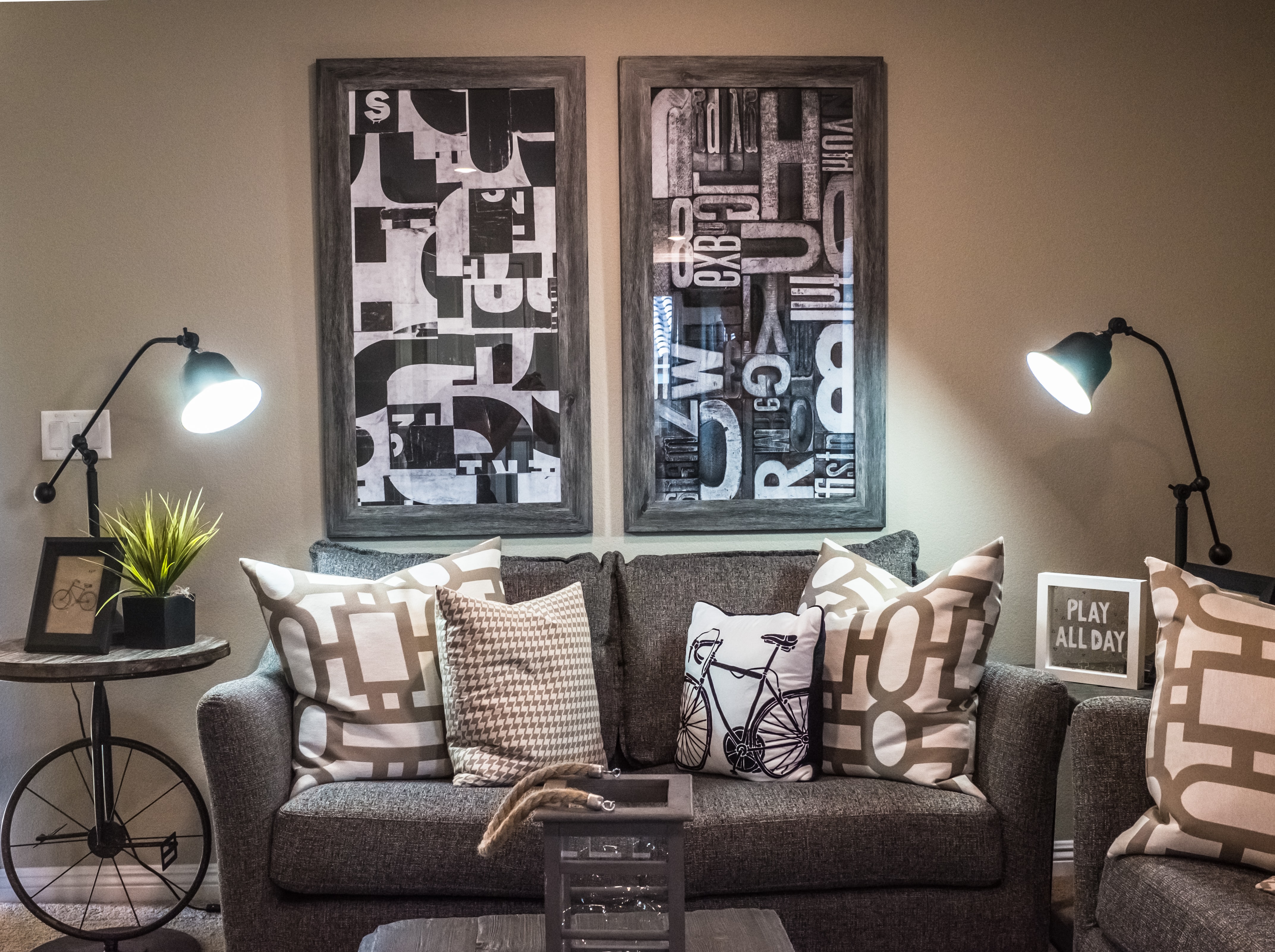 furnished corner of bicycle-themed room with gray couch, white throw pillows, lamp tables, and wall art
