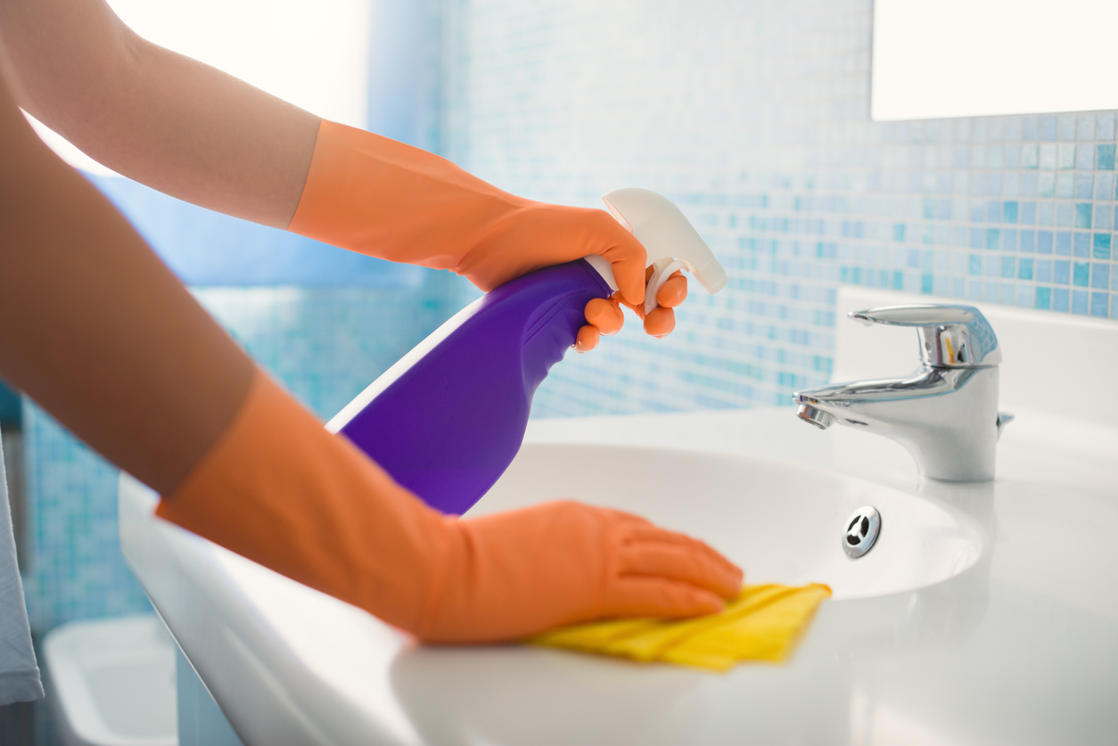 person in orange gloves cleans bathroom sink with purple cleaner and yellow towel. 
