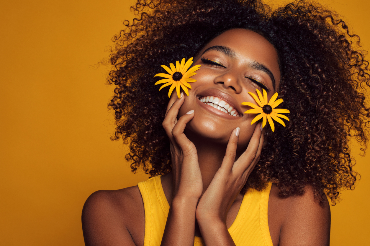 african american woman in yellow tank in front of yellow background smiles while holding two sunflowers. 