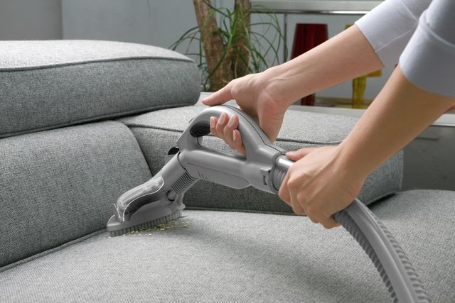 close view of a womans arms using a vacuum on a grey oversized chair to clean up chip crumbs