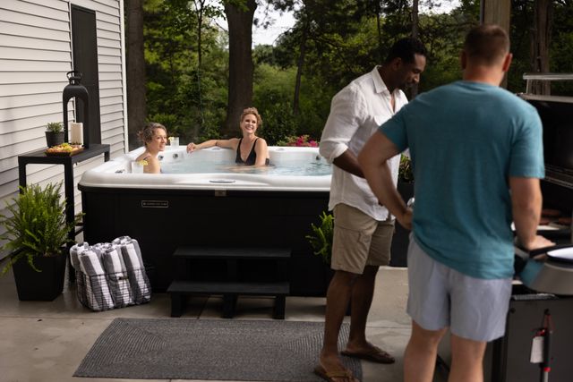 front view of two woman sitting in a Nordic hot tub while two men stand on the side and chat next to a grill