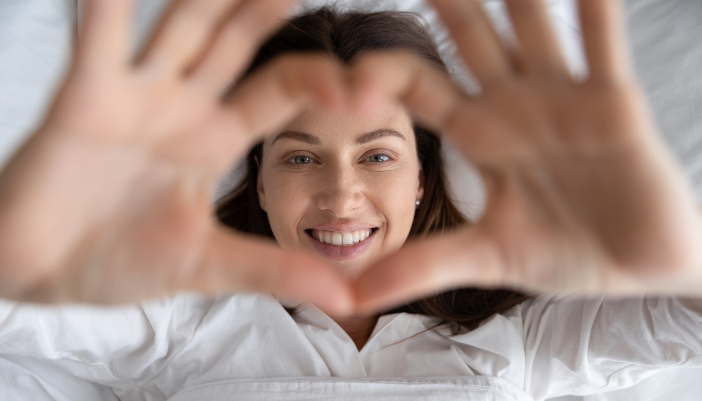 Woman smiling up at camera making a heart with her hands in bed