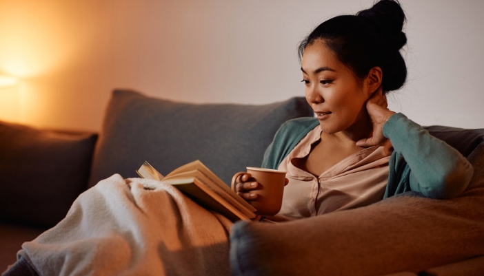 A woman in a warmly lit room drinks tea and reads a book.
