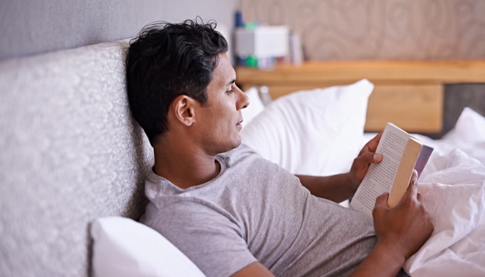 A man lays in bed at night and reads a thick paperback novel.