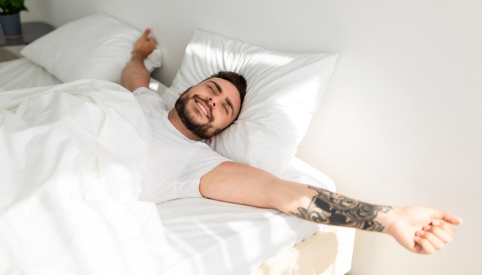 A tattooed man stretches and wakes up happily in his white bedsheets. 