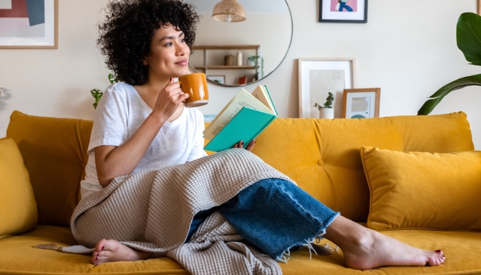 A woman sits on her couch with a book and a cup of coffee. 