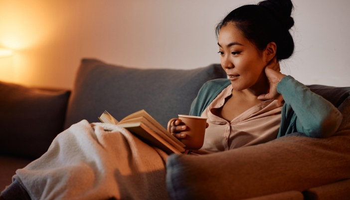 A woman sits on her couch with a book and a cup of tea.