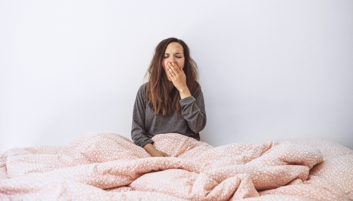 A girl in bed under a pink comforter and yawning.