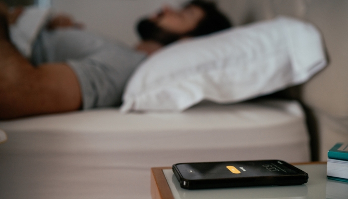 A man sleeps in his bed and an alarm is visible in the forefront.