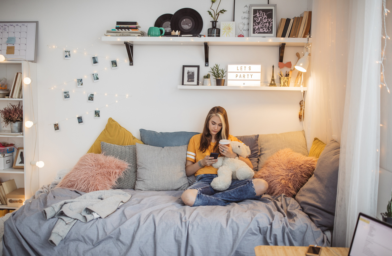 young woman sitting on college bed, holding teddy bear and using mobile phone