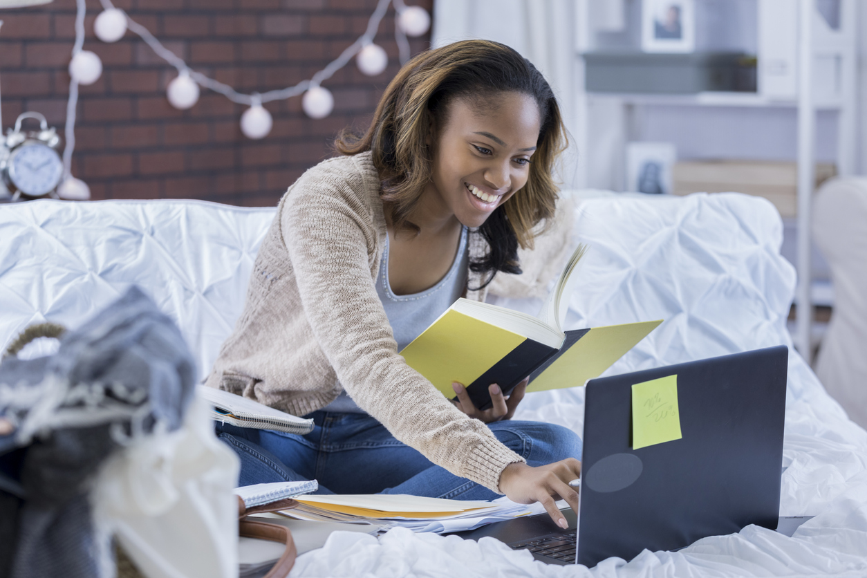 young woman sitting on college bed, holding teddy bear and using mobile phone