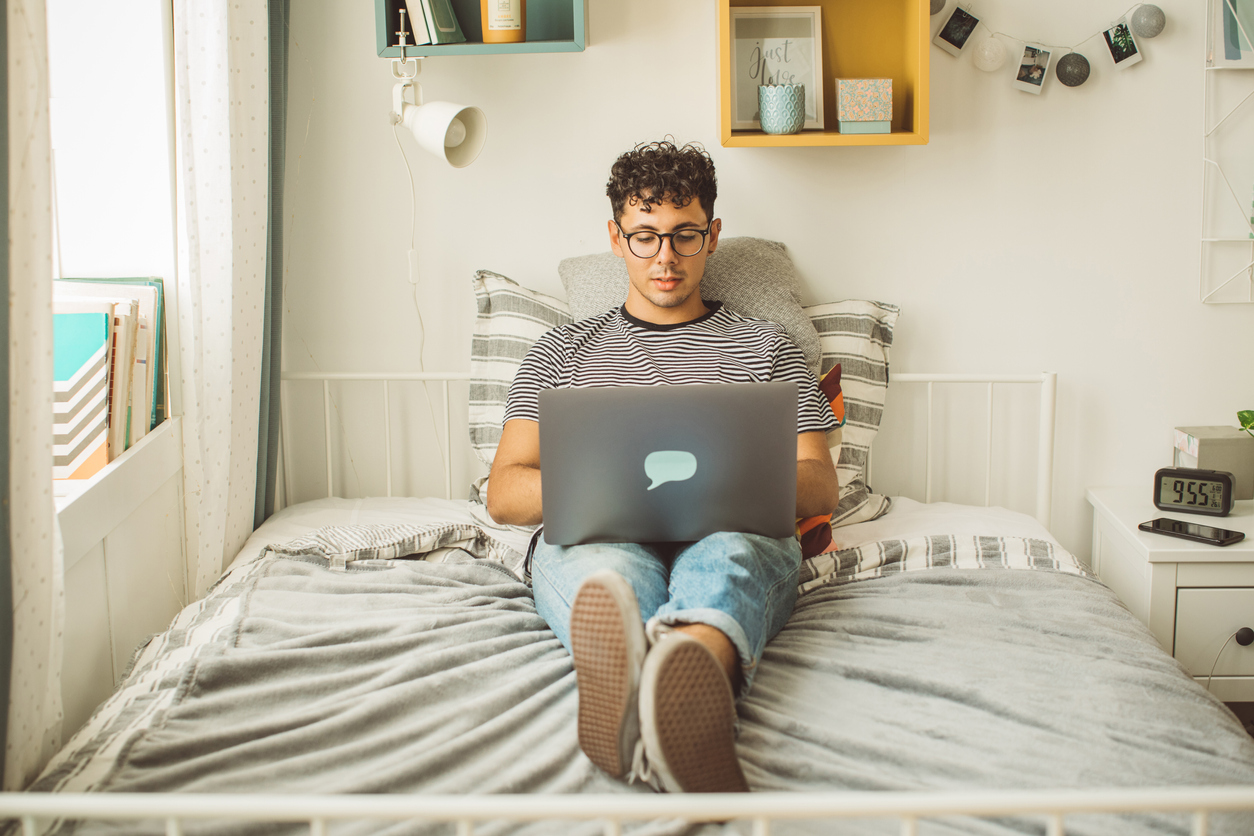 young man uses laptop while studying on college mattress