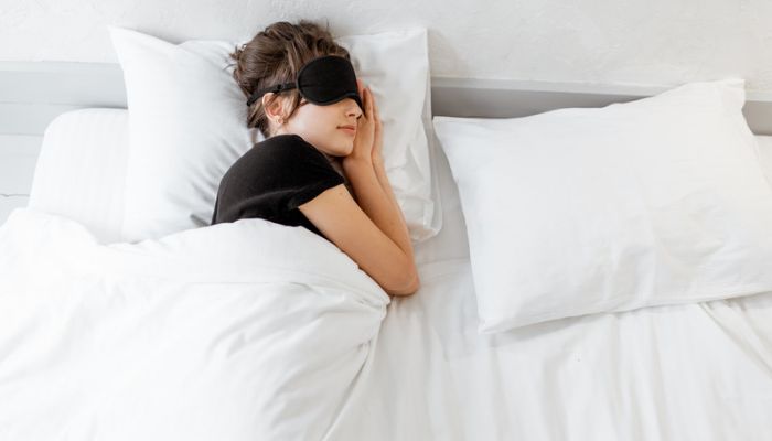 A woman lays in bed with a black sleep mask over her eyes