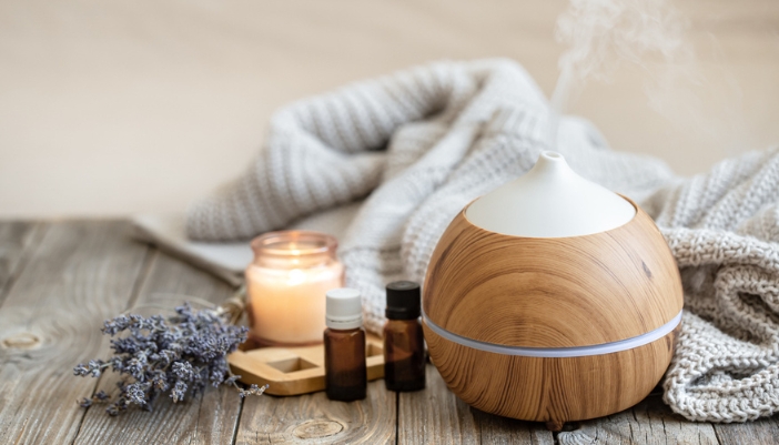 An essential oil diffuser sits on a wooden floor with a blanket and candle around it.