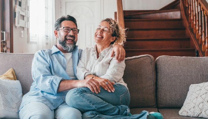 A mature couple sits on a gray couch and laughs together.