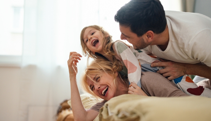A mother and father play-wrestle with their young daughter on a bed.