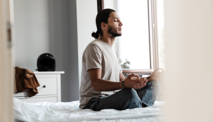 A man sits on his white made up bed and meditates calmly.
