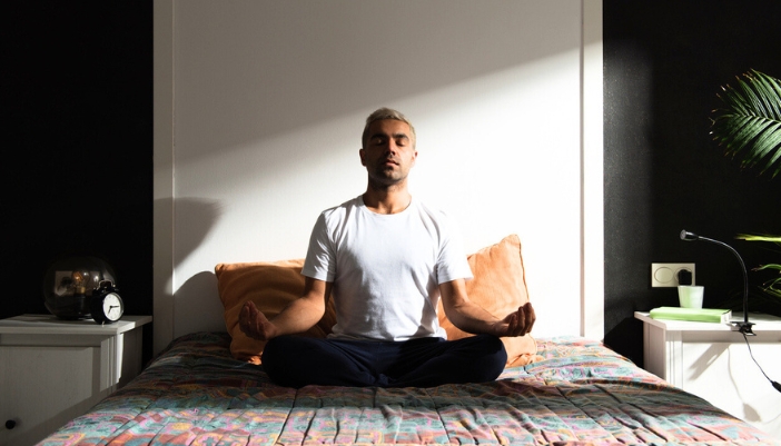 An ethnic man sits on a colorful bedspread while meditating calmly.