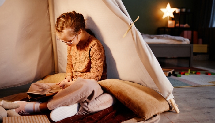 A young girl reads a book in a pop-up tent in her dimly lit bedroom.