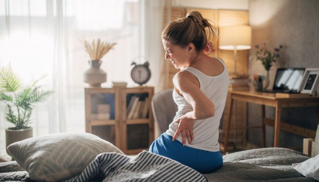 View of a woman scrunching her face and holding her back in pain on an old mattress in a modern bedroom