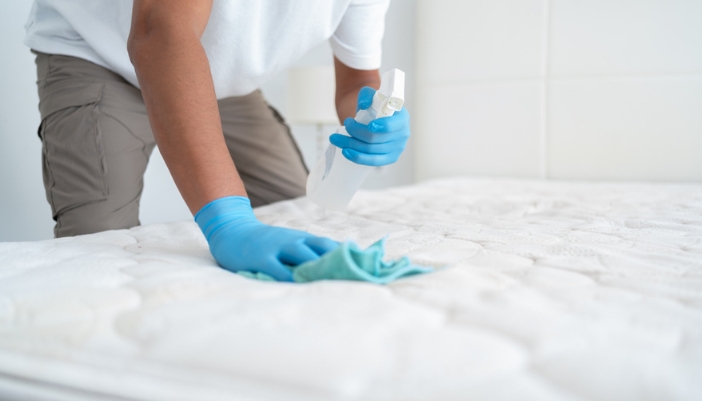 A man’s hand sprays cleaning solution on a mattress to scrub.