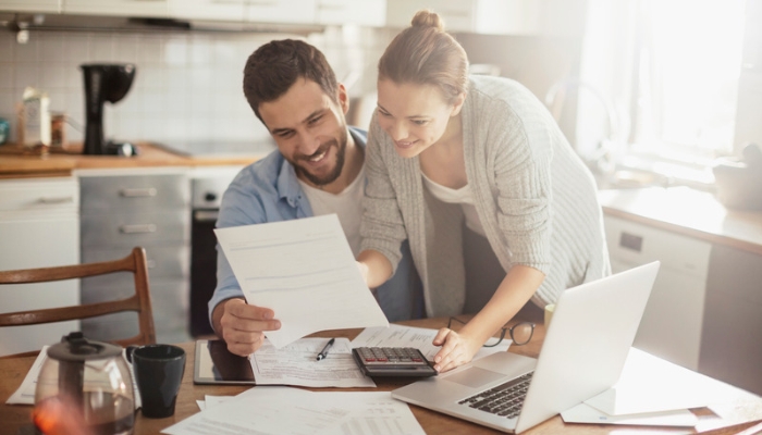 A couple sits with a computer and a laptop to work out a budget spreadsheet.