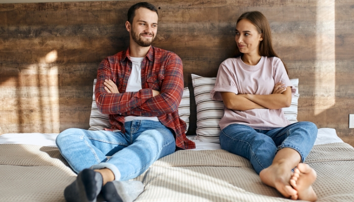 A couple sits on opposite sides of a bed with crossed arms and smirks. 