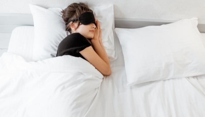 A woman with a black eyemask sleeps on a bed with white bedding.
