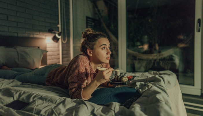 A woman lays on her bed late at night watching TV and eating sushi.