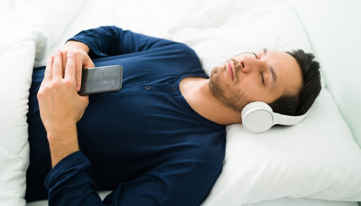 A man relaxes on his bed with over ear headphones.