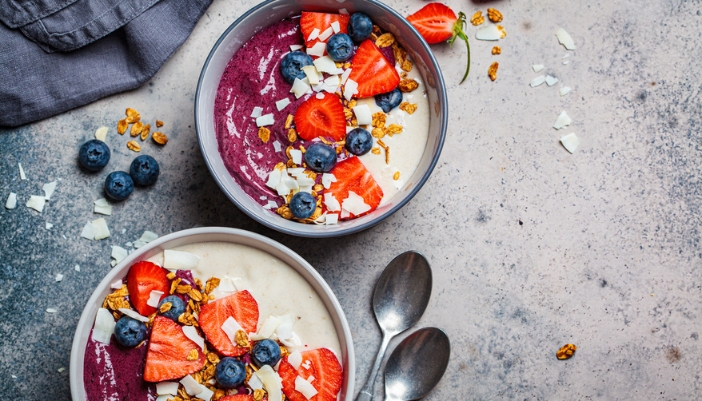 A pair of purple smoothie bowls rests on a background with fruit and granola.