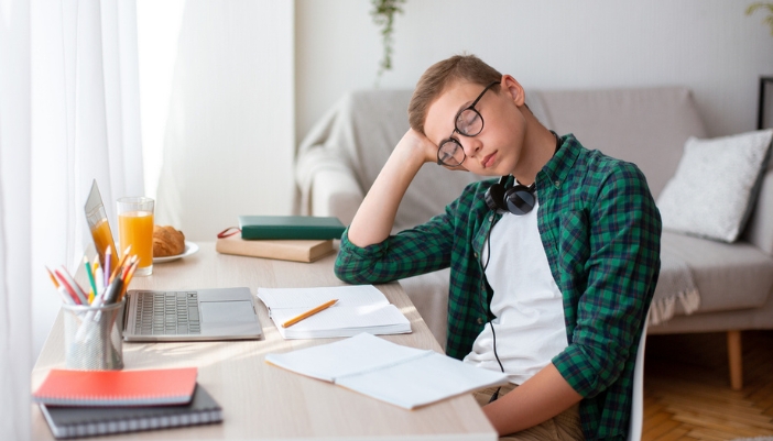 A teen sleeps at a desk with homework open in front of him.