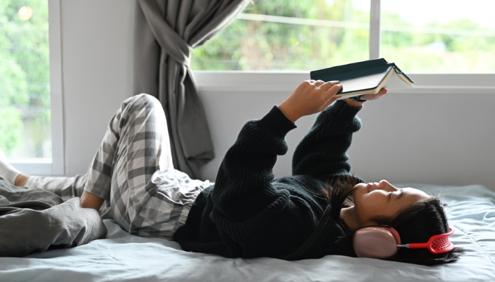 A teenage girl lays in bed and reads a book with headphones on.