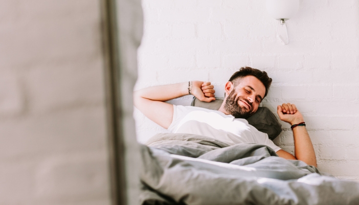 A happy man in a brightly lit bedroom lays down in his comfy bed.