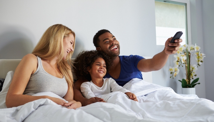 A mixed race family lays in their bed and chooses something to watch on TV. 
