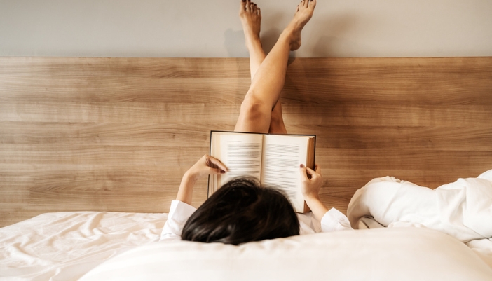 A woman lays on her bed with her feet up her headboard reading a book.