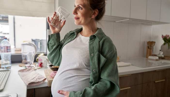 A young pregnant woman sips a glass of water as she prepares a meal.