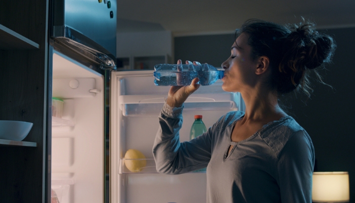 A woman chugs a water bottle in a dark kitchen at night.