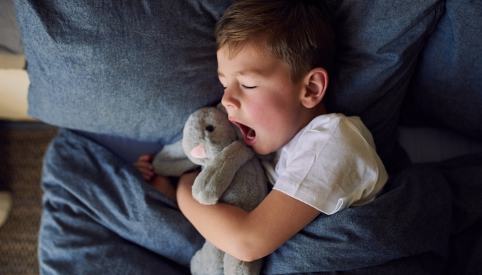 A young boy yawns in bed hugging a stuffed bunny.