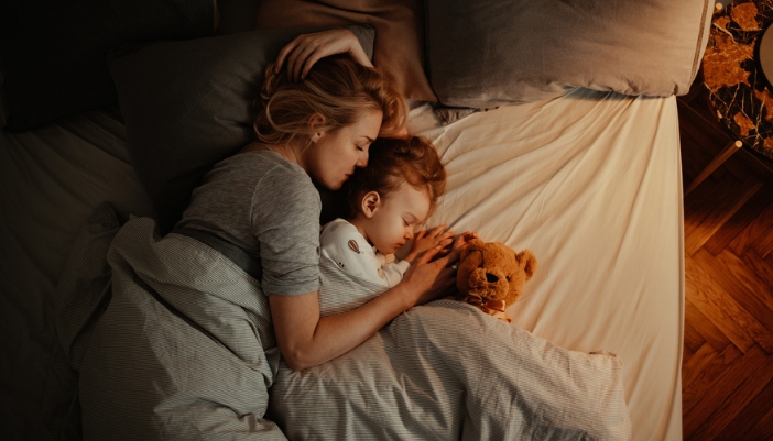 A mom and young girl sleep together in a warmly lit bedroom.