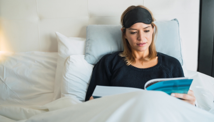 A woman lays in bed reading a book and wearing a black eye mask on her forehead.