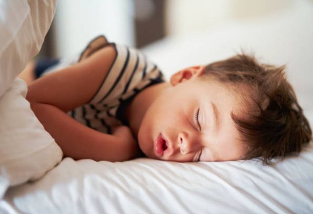 side view of a toddler in a striped shirt sleeping on a bed with a white comforter and sheets