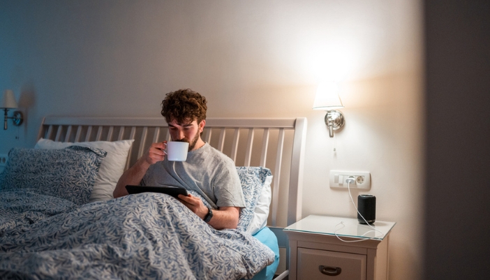 A man sits in a dimly lit room reading a book and drinking tea.