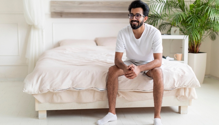 A well-rested man sits happily on the end of his bed.