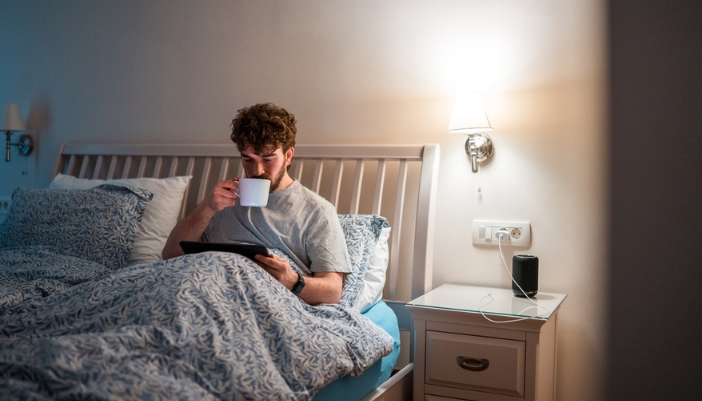 A man with curly brown hair lays in bed reading a tablet and drinking from a mug.
