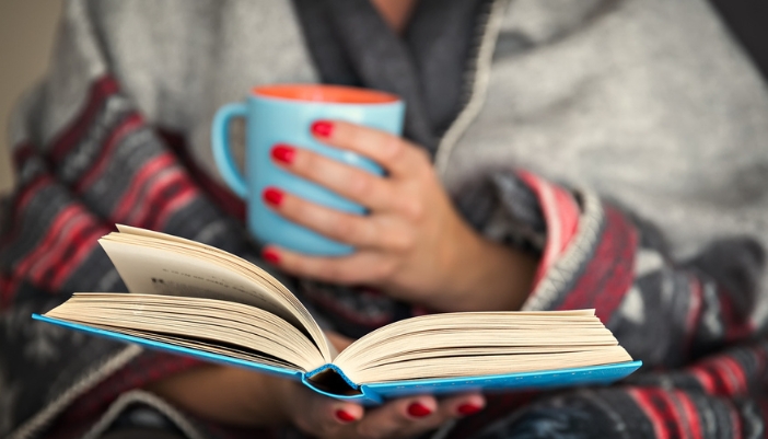 A woman wrapped up in a gray blanket, in the forefront is an open book and mug.