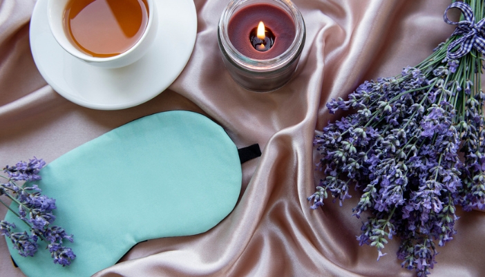 A lit candle, cup of tea, and blue night mask rest on pink satin next to lavender.