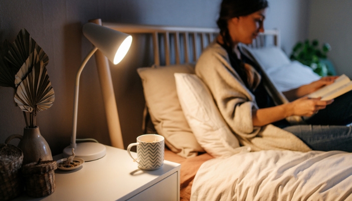 A woman lays in bed reading a book with a mug on the nightstand beside her.